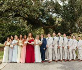 Wedding party posing under a large Louisiana tree, bridesmaids in pastel and red, groomsmen in light suits, lush greenery all around.