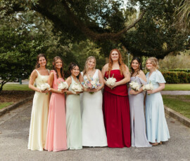 Seven women in colorful dresses hold bouquets and smile outdoors amid trees, celebrating a vibrant Louisiana wedding.