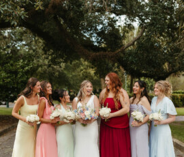 Seven women in colorful dresses hold bouquets and smile outdoors, capturing the joyful spirit of a Louisiana wedding.