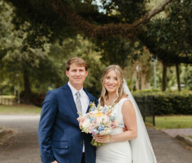 Bride in white dress and veil with groom in blue suit, smiling and holding pastel bouquet under large Louisiana tree.