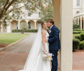 Bride in white gown with bouquet and groom in navy suit share a forehead kiss under a garden archway.