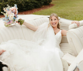 Smiling bride in white dress with pastel bouquet sits in white convertible, greenery around, on a sunny Gulf South wedding day.