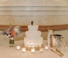 Elegant three-tiered white wedding cake with bride and groom topper, candles, flowers, champagne, and glasses on a round table.
