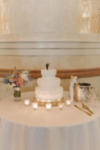 Elegant three-tiered white wedding cake with bride and groom topper, candles, flowers, champagne, and glasses on a round table.