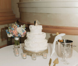 Four-tier white wedding cake with gold utensils, champagne, flowers, and candles on a table; elegant architectural background.