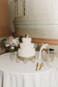 Four-tier white wedding cake with gold utensils, champagne, flowers, and candles on a table; elegant architectural background.