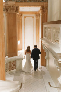 Bride and groom descend marble stairs in a grand, warmly lit hall with gold accents at a New Orleans wedding.