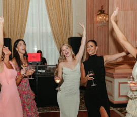 Five women in dresses celebrate with drinks at a wedding reception, smiling under elegant drapes and chandeliers.