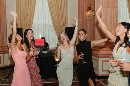 Five women in dresses celebrate with drinks at a wedding reception, smiling under elegant drapes and chandeliers.