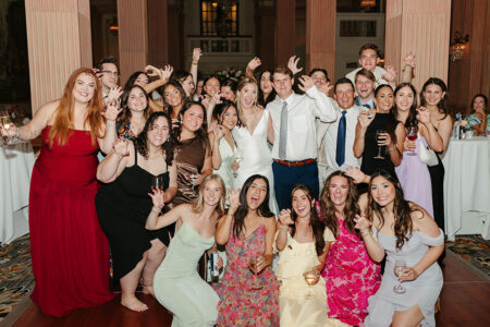 Formal wedding guests pose indoors with drinks, making playful claw gestures and smiling at a joyful Gulf South celebration.