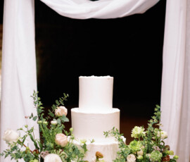 Elegant two-tiered white cake with green and white flowers, under draped white fabric, ideal for a Louisiana wedding.