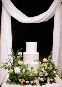 Three-tier white wedding cake surrounded by lush floral arrangements of green and blush flowers, displayed under soft white draping at a romantic reception setup.