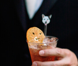 Person in dark suit holding cocktail with ice, dried orange slice, and cartoon dog stir stick—ideal for a Louisiana wedding.