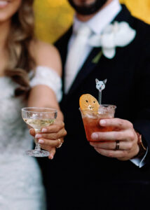 Close-up of bride and groom holding their signature wedding cocktails, including a champagne coupe and a custom drink with a dog-shaped stir stick and dried citrus garnish, celebrating in elegant wedding attire