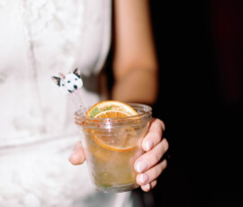 A person in a white strapless dress holds a cocktail with an orange slice and dog stirrer at a festive New Orleans wedding.