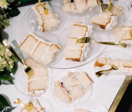 White layer cake slices with gold forks on glass plates, set on a white table with green and white floral decor.
