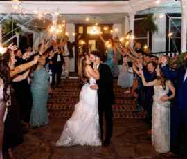 Bride and groom kiss at night, surrounded by cheering guests with sparklers outside a decorated Louisiana wedding venue.