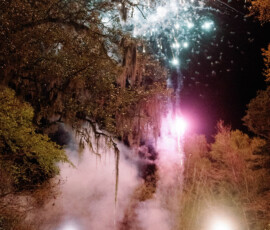 Red car under trees at night with smoke and fireworks in the sky, evoking a festive New Orleans wedding atmosphere.