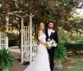 Bride in white gown and groom in black suit smile under flowering archway, holding white bouquet at classic Louisiana wedding.