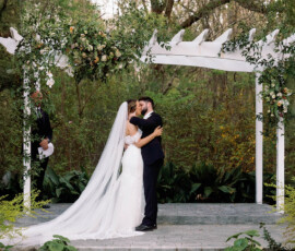 Bride and groom kiss beneath a white, flower-adorned pergola at their Louisiana outdoor wedding, surrounded by greenery.