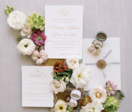 Elegant wedding invitation suite with cream and blush florals, gold script, rings, wax seal, and pearl earrings on linen.
