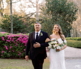Bride in white gown with bouquet walks outdoors, arm-in-arm with man in dark suit; pink flowers and greenery behind.
