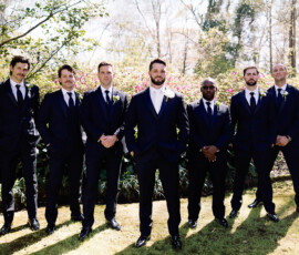Six men in dark suits pose on grass at a Louisiana wedding, with the groom standing slightly ahead in sunlight and greenery.