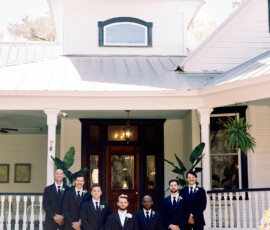 Seven men in suits pose on brick steps before a classic white Louisiana house with porch, columns, and tall windows.