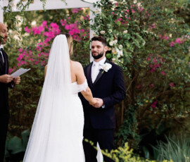 Bride and groom face each other under a white pergola at an outdoor Louisiana wedding, surrounded by greenery and pink flowers.