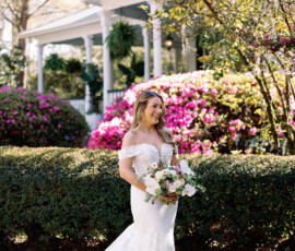 Bride in off-shoulder white gown with bouquet stands in sunny garden, white house behind, New Orleans wedding elegance.