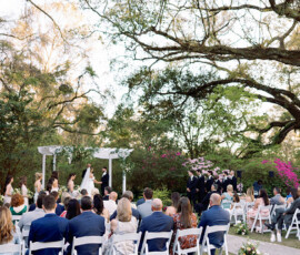 Gulf South outdoor wedding with guests on white chairs, couple under a pergola, lush greenery, flowers, and large arching trees.
