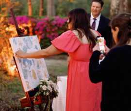 Woman in coral dress paints at outdoor wedding as guest takes photo; man in suit and flowers in background.