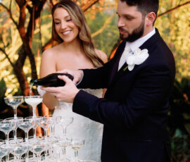 Bride and groom pour champagne into a glass tower at their outdoor wedding, surrounded by greenery and white flowers.
