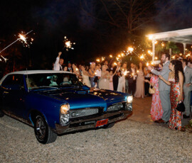 Blue classic car passes a crowd with sparklers at night as people in formal wear celebrate outside a house, New Orleans style.
