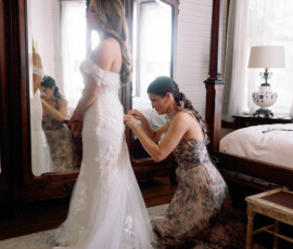 A woman kneels, fastening a bride’s lacy gown in a softly lit bedroom with an ornate mirror reflecting the Gulf South wedding moment.