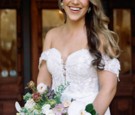 Smiling bride in an off-shoulder white wedding dress holding flowers stands before wooden doors at her Louisiana wedding.
