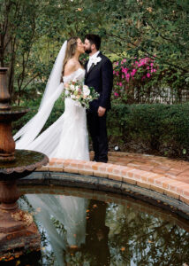 “Bride and groom share a kiss beside a garden fountain, with the bride in an off-the-shoulder lace gown and long veil holding a romantic bouquet, surrounded by lush greenery and blooming pink flowers.”
