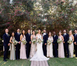 Wedding party in front of blooming trees, with bride, groom, bridesmaids in light dresses, and groomsmen in dark suits smiling.
