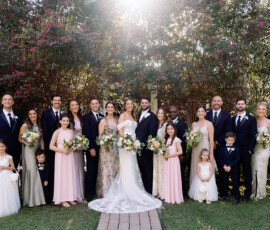 Outdoor wedding party poses and smiles in sunlight, with bridal party in classic attire, beneath leafy trees.