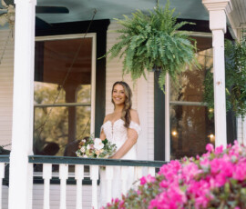 Smiling woman in white dress on porch with bouquet; pink flowers, green ferns, and white house in background.