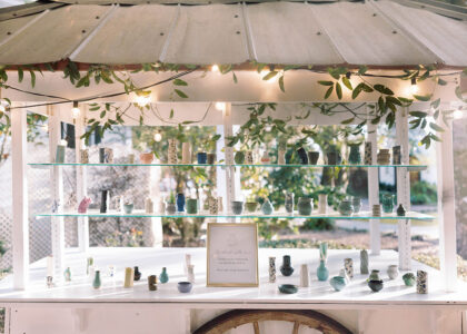 Display of handcrafted ceramic wedding favors arranged on glass shelves of a white cart decorated with greenery and string lights at an outdoor reception.