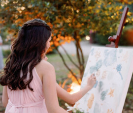 Woman in pink dress paints outdoors on easel, surrounded by greenery and warm lighting at a Louisiana wedding.