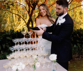 Bride and groom smile outdoors by a champagne tower, pouring champagne, with flowers and greenery creating a romantic setting.