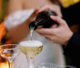 Champagne pours into a glass tower with overflow; woman in white dress blurred behind, evoking Gulf South wedding elegance.