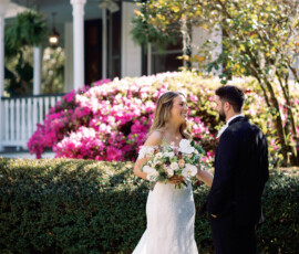 Bride and groom smiling outdoors by a house with pink flowers at their New Orleans wedding; she holds a bouquet.