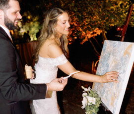 Bride and groom in wedding attire smile while painting together at their nighttime outdoor Louisiana wedding reception.