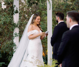 Bride in white reads vows to groom under floral arch at outdoor Louisiana wedding, groomsman in foreground.