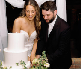Bride and groom cut white tiered wedding cake amid flowers and greenery under white drapery at a Louisiana wedding.