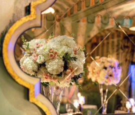 White and pale pink flowers in a glass vase reflected in an ornate gold mirror, with candles and elegant decor behind.