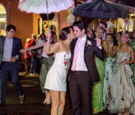 Formally dressed couple with umbrellas stands together at a lively Louisiana wedding, surrounded by cheering friends at night.